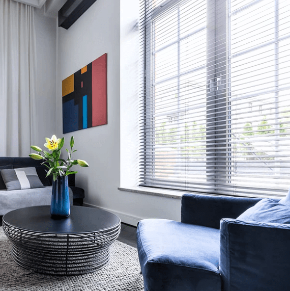 Silvriga aluminiumpersienner i lounge vid stort fönster, modern soffa och soffbord på ljus matta