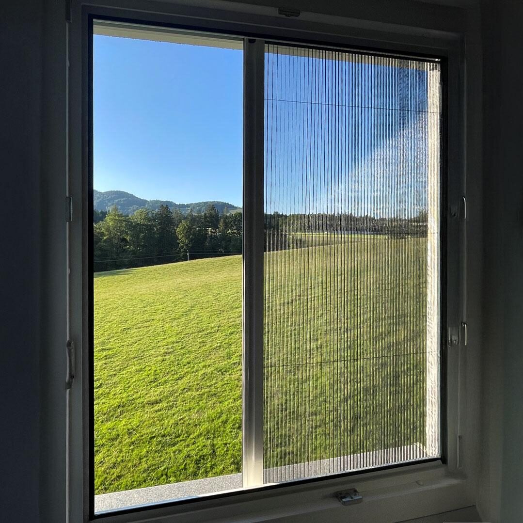 Plissémyggnät vid fönster – insektsfritt, ljusinsläpp och klar utsikt över äng.